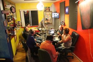Photograph of 6 people sitting around a table with microphones and headphones in the middle of a recording that may or may not be a podcast.