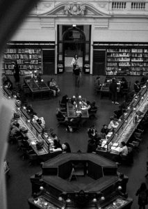 Black and white photograph of what may be a library reading room. The picture is taken from a second floor or balcony, providing a bird's eye perspective. There are several patrons reading, studying, and writing at two diagonal tables, sofas, and desks, as well as books on shelves along the wall.