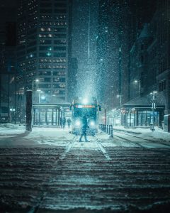 Gorgeous night time photo of a train in an undisclosed downtown area of a city in a snow storm. A pedestrian is walking in front of the train, while the street lights provide an eerie glow, highlighting this lovely and disturbing image.