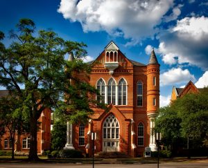 Photograph of a red-brick university building, with cathedral windows and a slim tower. It is framed by two trees and a blue sky with aesthetically fluffy clouds.