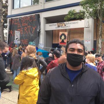 Person stands with crowd in background of Seattle Police Department Precinct during Black Lives Matter protests following the police killing of George Floyd