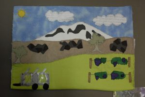 Image of the completed Midterm Project. It includes a mountain made of fabric, a dry hillside made of fabric with basalt rocks and sagebrush of fabric, a green agricultral slope of fabric with two rows of trellised grapes, a trailer with wheels and silver strips on top of it representing solar panels, a yellow sun in the top left corner, and two clouds cut from notes taken in college classes.
