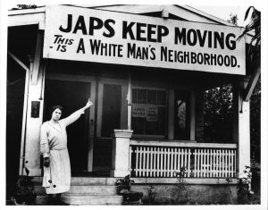 Photograph of women standing by a house pointing to a sign that says "JAPS KEEP MOVING THIS IS A WHITE MAN'S NEIGHBORHOOD."
