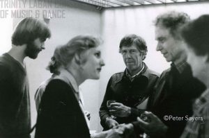 Five people are pictured having a discussion. The middle person is looking at the camera, and the two to each side of him are blurry. This middle person is John Cage.
