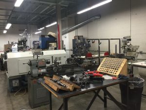 A picture of a community makerspace. Many tools atop a table are in the foreground, and various machines for fabrication are in the background. The room is brightly lit by overhead lights, and the walls are concrete blocks painted white.