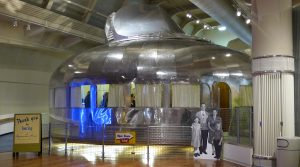 A shining metal circular building made of welded metal panels. Windows with curtains wrap around the middle of the form. The building is within a much larger museum.