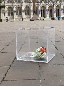Crumpled ball of grid-ruled engineering paper inside a transparent acrylic glass cube on a brick floor outside in front of a wide college-gothic building.