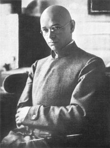 Black and white portrait of bald man wearing glasses, arms crossed, and looking down slightly.