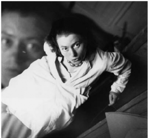 Double-exposed self-portrait photograph of woman in trench coat sitting on a chair and looking upwards at camera, with a secondary exposure of her face dominating the upper-left corner of the photo.