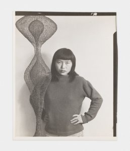 A photo of Ruth Asawa standing in front of a hanging sculpture