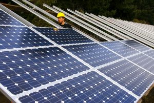 A man in a yellow hard hat stands behind many photovoltaic panels raised from the ground on an angled structure.