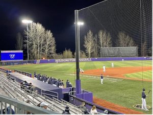 Alt text: This photo shows a baseball game at the University of Washington. The game is happening at night. You can see the stands, and there are a few spectators.
