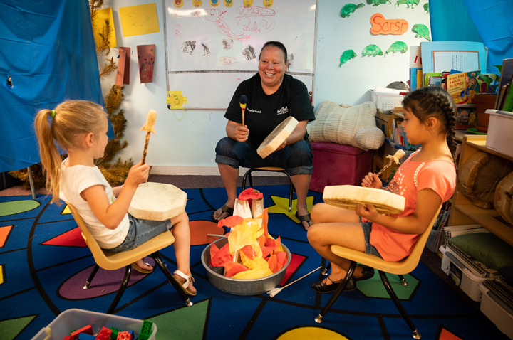 educator at a Salish Language Immersion Program seated in the dramatic play area of her classroom with two children. They're seated around a fire playing drums.