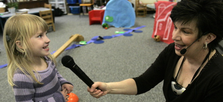 Image depicting a classroom with an audiologist holding out a microphone to and speaking with a child.