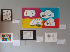 Five examples of student art on a grey cinderblock wall. Each piece of art has a title, artist name, and artist statement on a small bordered square next to the art. Images within the art include: indigenous women that have disappeared, a large four "bubbled" piece that follows the life of a male with the phrase" men are taught to bottle up their feelings", an outline of a head with a cloud and rain inside it that is filling the shape up with water, puzzle pieces with famous cartoon characters, and an image divided into four squares that shows someone struggling with their dark thoughts and then finding ways to express those thoughts that are healthy such as creating art or meditating.