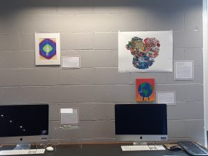 Three examples of student art on a grey cinderblock wall and one piece of student art on a clear pedestal. Each piece of art has a title, artist name, and artist statement on a small bordered square next to the art. Images within the art include: a person surrounded by colored rings, a collage of yellow, red, and blue images cut from magazines, the earth dripping blue on a background of orange. The 3-D object is a phone with a tennis net and court on top of it.