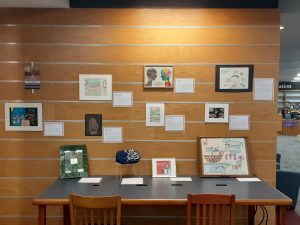 Twelve examples of student art on a wooden wall and on a large table. Each piece of art has a title, artist name, and artist statement on a small bordered square next to the art.