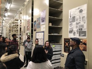 a group of people are gathered in the aisle of a museum collection hallway, watching a person in the center who is speaking. museum artifacts can be seen on the shelves surrounding the people.
