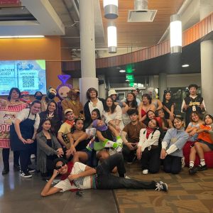 This image highlights the Sayaw (Filipino folk dance team) dressed up in costumes as several characters (Buzz and Woody from Toy Story, Scooby, and so much more). They are all gathered in the lobby of the Samuel E. Kelly Ethnic Cultural Center (the ECC) after their Halloween dance practice. Some are standing, some are crouching, one member is even posing on the floor.