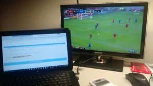 On a college dorm desk, the computer on left shows calculus homework. On the right side, there is a television screen playing a soccer game.