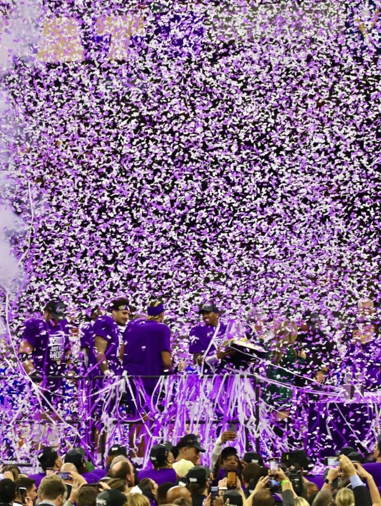 Image of confetti showering the football field at Caesars Superdome in New Orleans, Louisiana. The vibrant purple confetti celebrates University of Washington's 37-31 victory over University of Texas, with fans in purple attire cheering in the background.