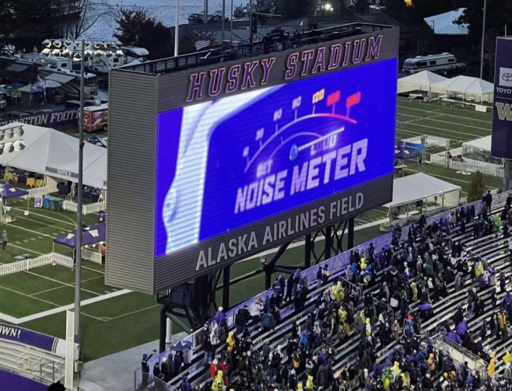 The photo shows a large screen at Husky Stadium in Seattle, Washington. The screen is displaying a noise meter, which is measuring the crowd noise in decibels. The meter is currently reading 105 decibels.