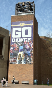 Image of a banner hanging on the UW clocktower outside Kane Hall. There is aperson dressed as UW Husky Mascot 'Dubs' in the stands with other fans at a UW football game