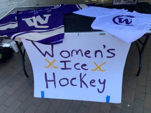 Informational image showing a table with UW jersey and shirt displayed on top and a hand painted poster hanging off the front of the table with Women's Ice Hockey