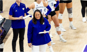 A middle-aged woman stands in the foreground of the photo on the side of a volleyball court. 4 female volleyball players in knee pads and jersey's stand behind her. A young man with a clipboard and blue shirt stands next to them.