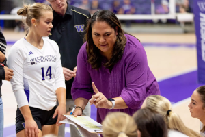 A woman in purple holding a clipboard talks to youngwomen in volleyball uniforms on the side of a court with a purple and wood floor.