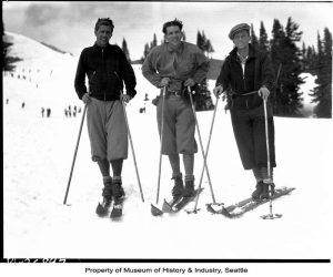 A black and white photo showing three men on skis holding ski poles. They are dressed similarly in pants, coats, and hats. They look at the camera smiling. They are on a white, snow-covered hill with dark evergreen trees in the background. Other people spot the hill behind them int he background of the photograph.