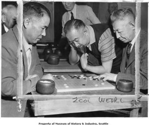 A black and white photograph showing 5 men leaning over a game board with black and white stones.