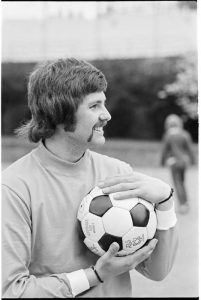 A historic Seattle soccer player holding a soccer ball, smiling during a casual moment, with a blurred field and figure in the background.