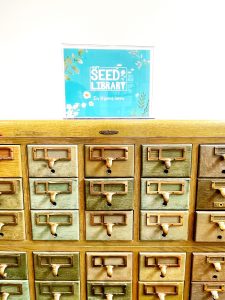 A vintage card catalog with shiny brass knobs. On top is a sign that reads, "Seed Library in Progress."