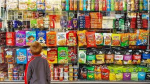 Blond child standing in front of a tall shelf full of chips, candy, cereral and other processed foods